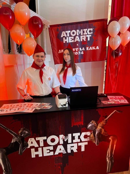 Промостойка игры Atomic Heart с ведущими в новогодних шапках, воздушными шарами и ноутбуком для регистрации