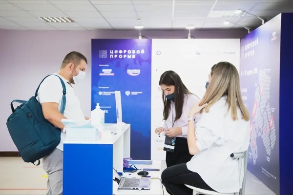Мужчина с рюкзаком получает бейдж и антисептик у стойки форума «Цифровой прорыв», сотрудницы в масках