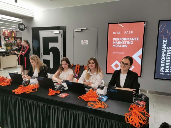 Четыре сотрудницы печатают бейджи для Performance Marketing Moscow за столом с оранжевыми лентами