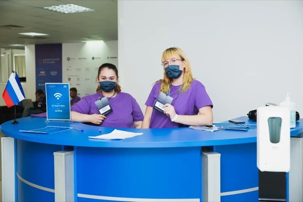 Две сотрудницы в фиолетовых футболках и масках стоят за синей круглой стойкой с флагом России