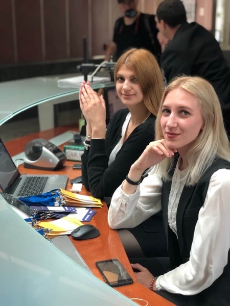 Две сотрудницы улыбаются за стойкой регистрации с ноутбуком и стопками бейджей на лентах