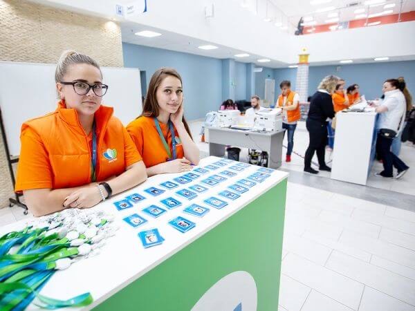 Две сотрудницы в оранжевых жилетах сидят за зелёной стойкой с разноцветными бейджами-значками
